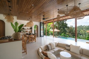 Un espace de vie moderne avec un plafond en bois, des luminaires en fibres naturelles, une grande plante, un ensemble de salle à manger et une vue sur une piscine.