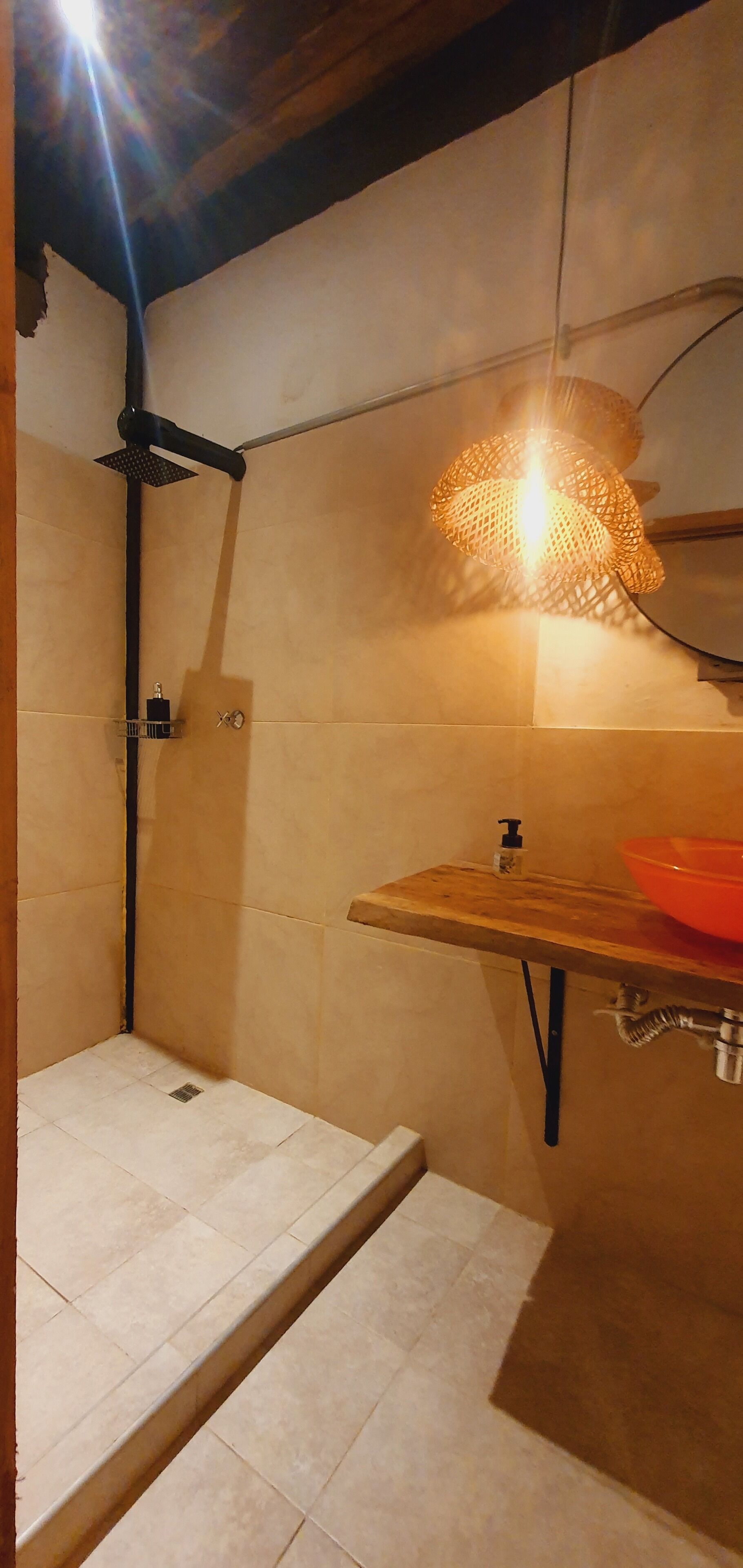 Un baño con ducha, un estante de madera con un lavabo rojo, y una lámpara colgante.