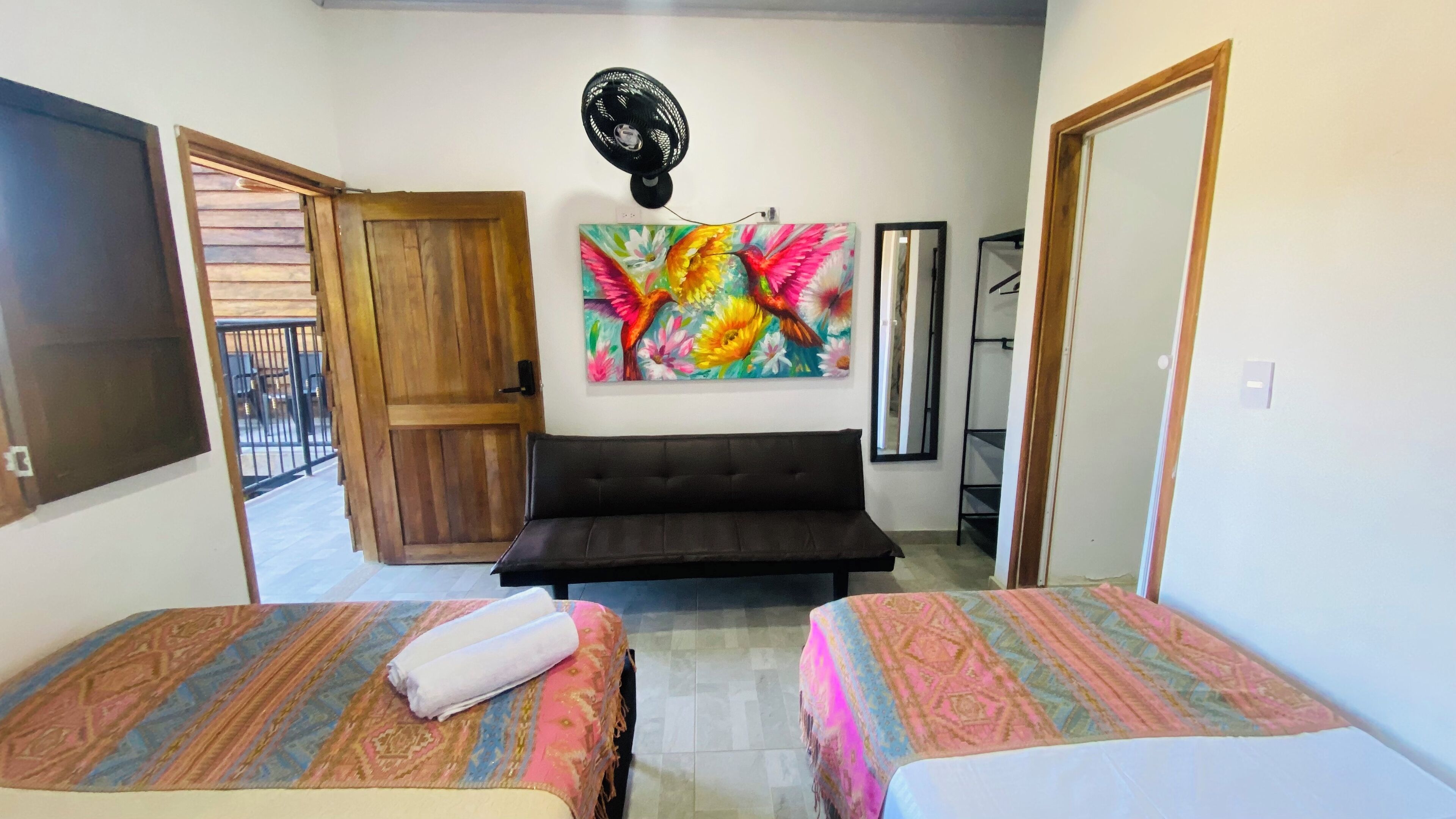 A hotel room with two beds, a sofa, a wall-mounted fan, and a colorful painting.