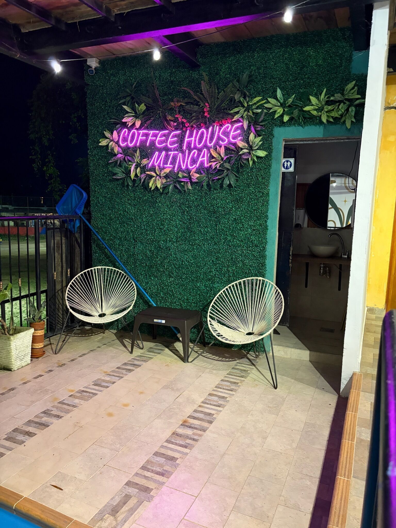 An outdoor seating area with two white chairs and a neon sign that reads 'Coffee House Minca.'