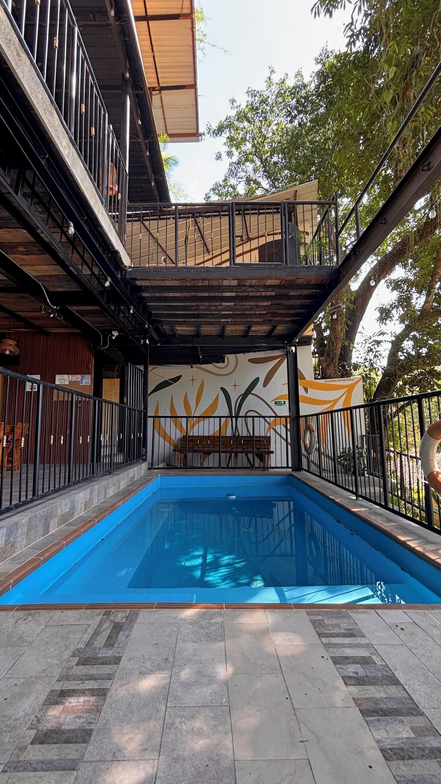 A swimming pool with a tiled deck, surrounded by a metal fence and a covered area with a mural.