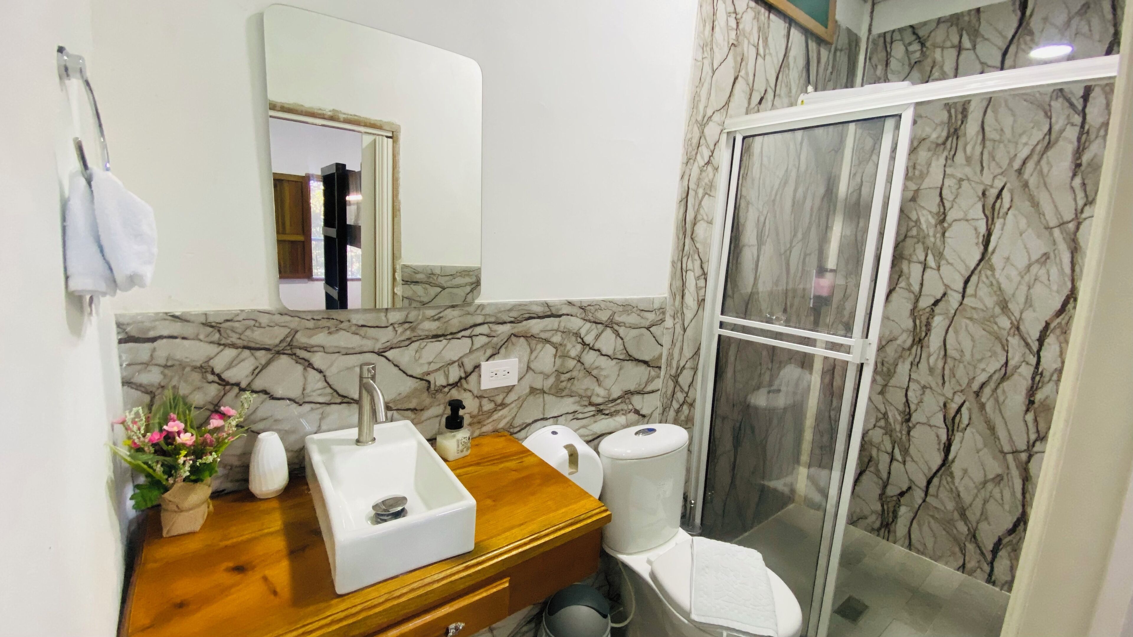 A bathroom with a marble backsplash, a wooden vanity, a sink, a mirror, a toilet, and a glass shower enclosure.