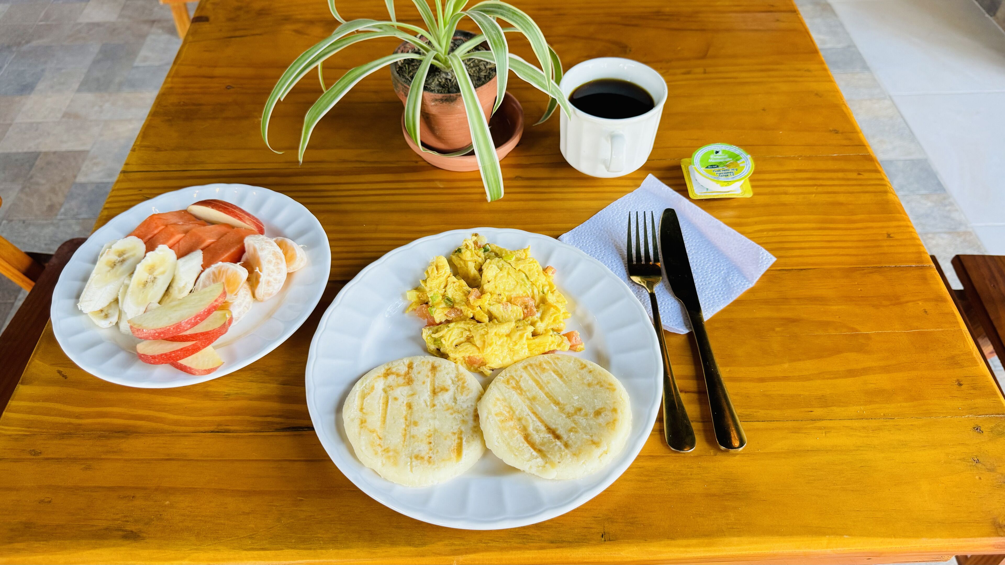 Une table en bois avec des assiettes de petit-déjeuner, une tasse de café, une plante et un petit pot de yaourt.