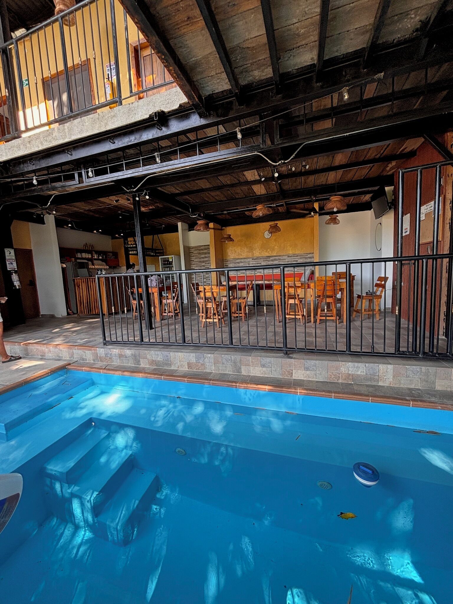 A swimming pool with a metal fence, wooden tables and chairs, and a covered patio area with a rustic ceiling.