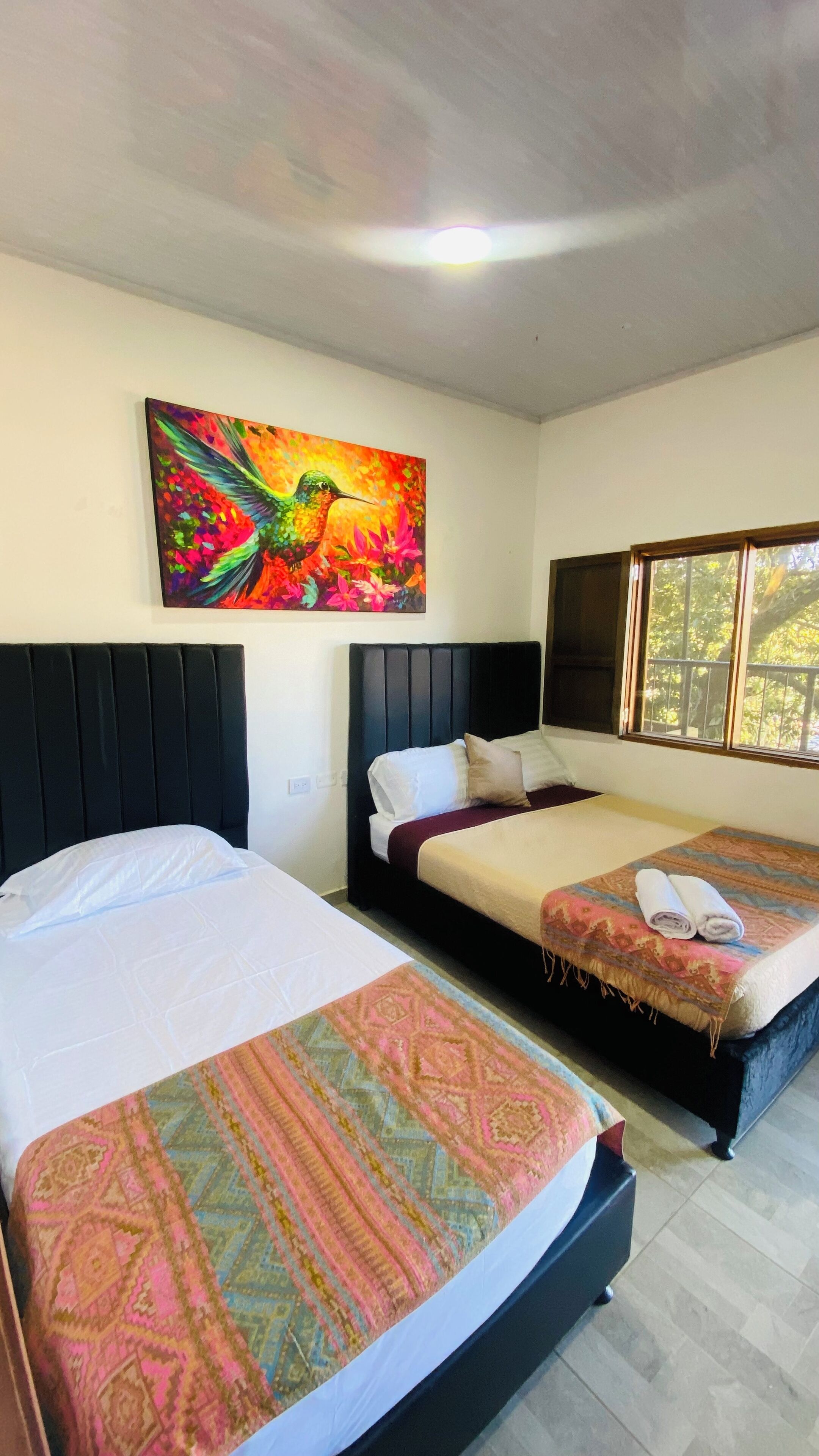 A hotel room with two beds, a large painting of a hummingbird, and a window with a view of trees.
