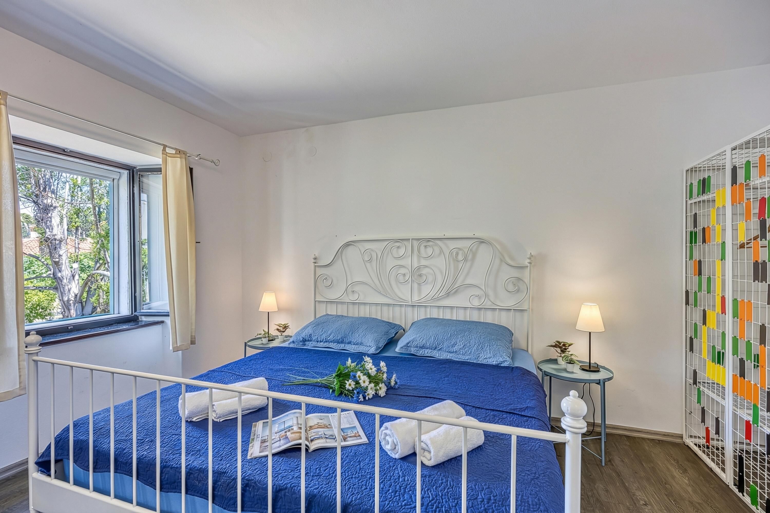 Ein Schlafzimmer mit einem Bett, zwei Nachttischen, einem Fenster mit Vorhängen, einer Wandverkleidung und Blick auf Bäume.