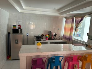 A kitchen with white tiles, a refrigerator, a microwave, a stove, and a sink. There are colorful plastic chairs and a small yellow duck figurine on the counter. - Secretos del Sol (Acapulco)