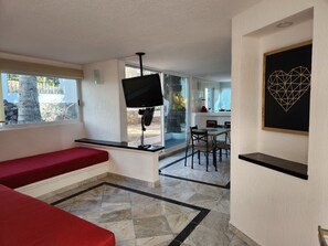 A modern room with a red bench, a flat-screen TV, a dining area with a table and chairs, and a geometric art piece on the wall. - Secretos del Sol (Acapulco)