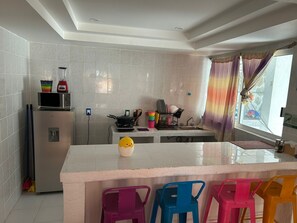 Una cocina con azulejos blancos, un refrigerador, un microondas, una estufa y un lavamanos. Hay sillas de plástico de colores y una pequeña figura de patito amarillo sobre la encimera.