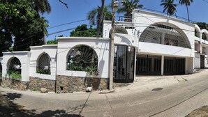 A white building with arched windows and a stone base, located at a street corner with palm trees and a clear sky. - Secretos del Sol (Acapulco)