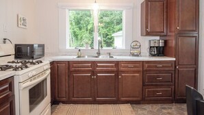A kitchen with wooden cabinets, a white stove, a microwave, a sink, and a window with a view of greenery.