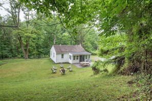 A small white house with a covered patio and outdoor seating area surrounded by dense trees. - Bright Cottage Retreat in Central PA (Mcveytown)