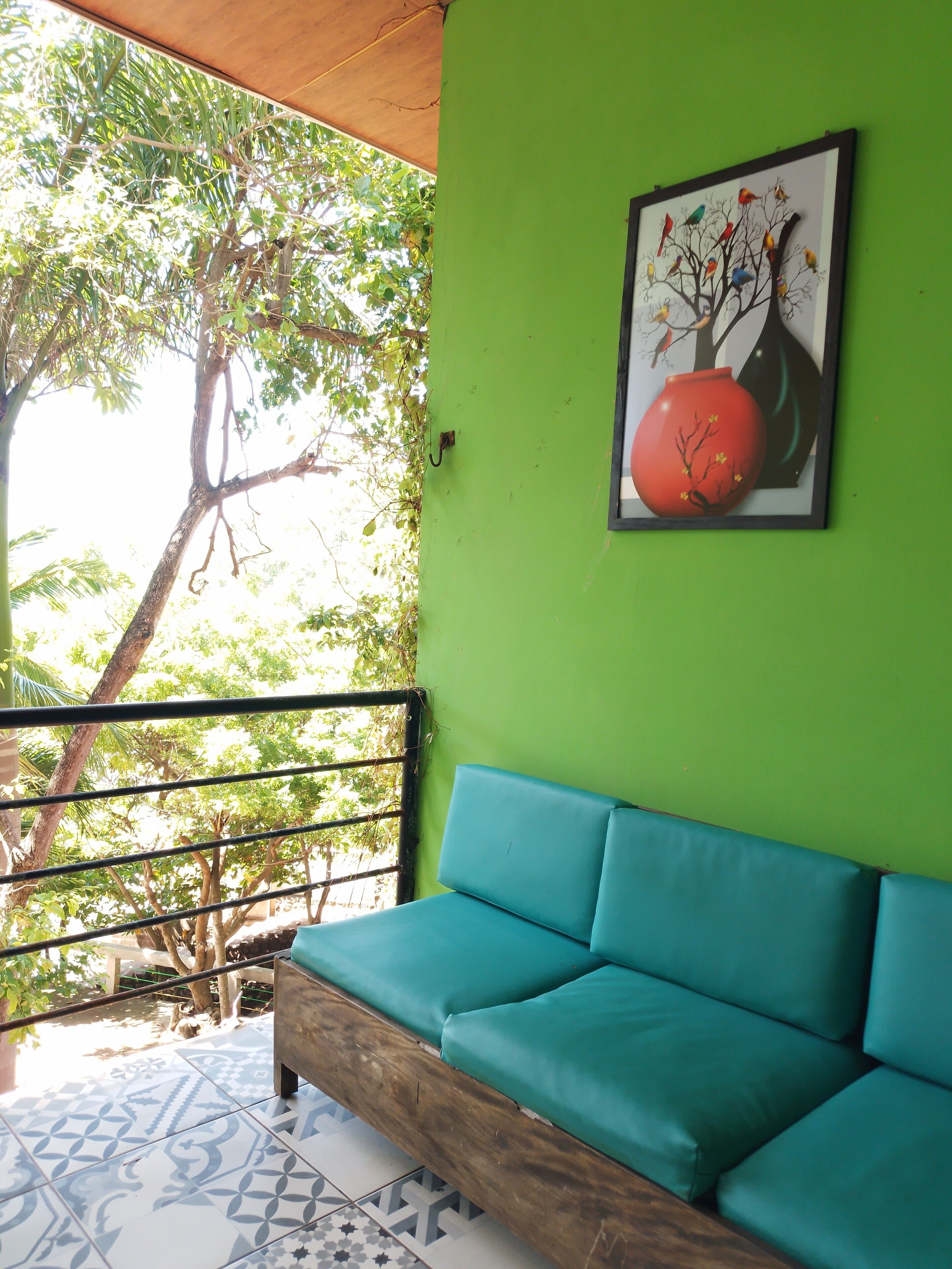 Una pared verde con un cuadro enmarcado de un árbol y un jarrón, un sofá color turquesa, y un balcón con barandilla y árboles al fondo.