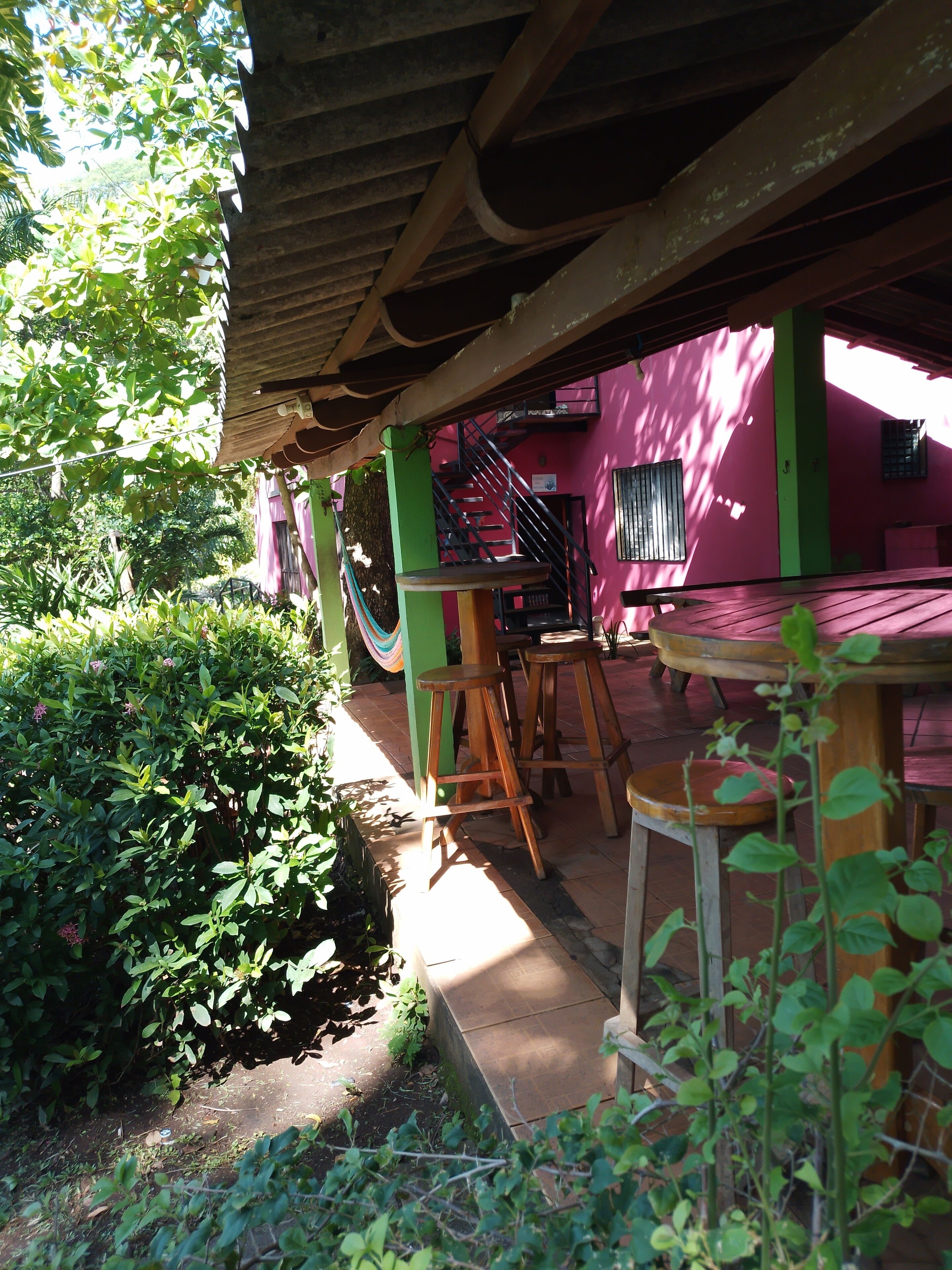 A covered outdoor seating area with wooden stools and a table, surrounded by greenery and a pink building with a staircase.