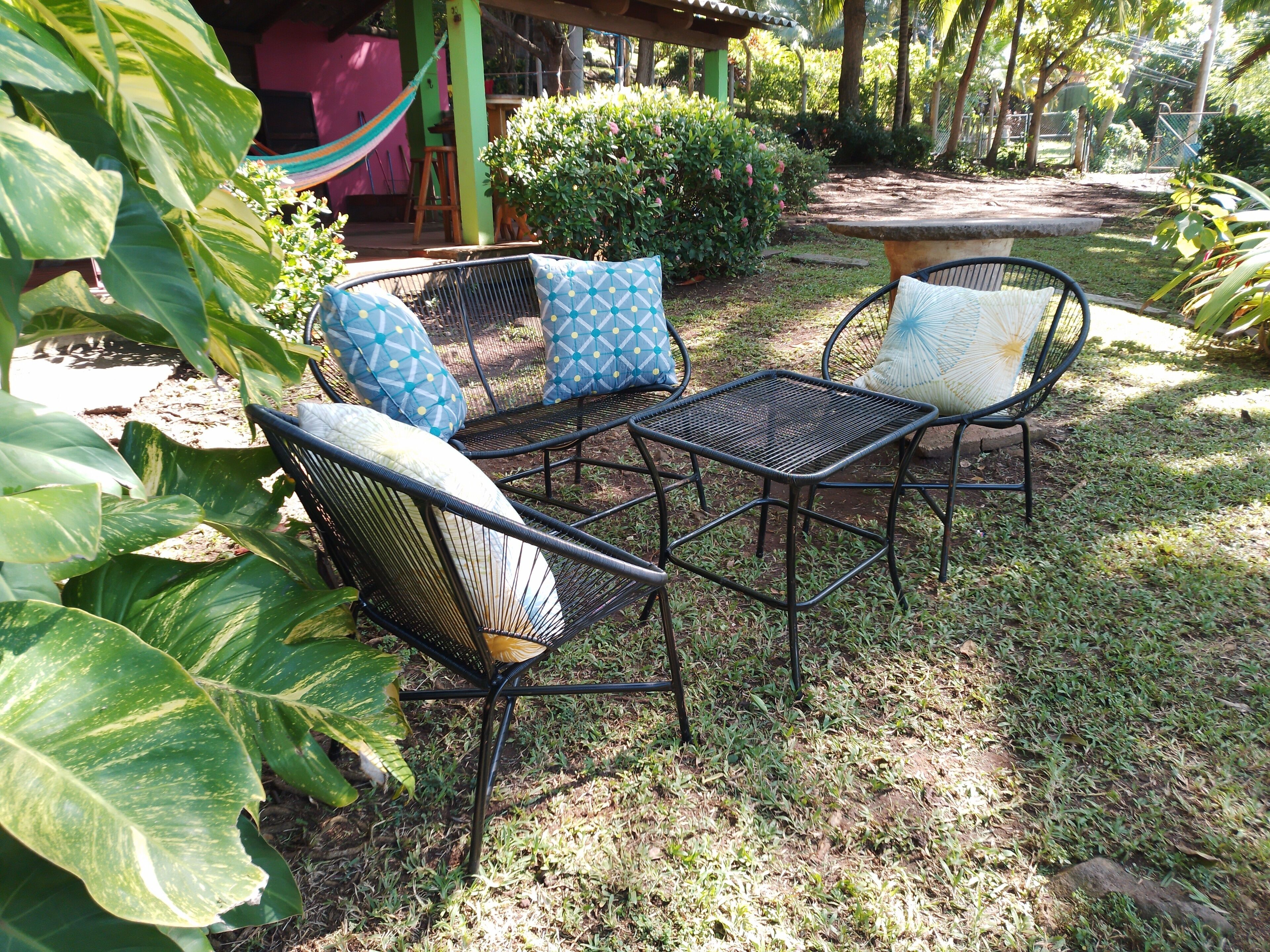 An outdoor seating area with metal chairs and cushions, surrounded by lush greenery and a hammock.