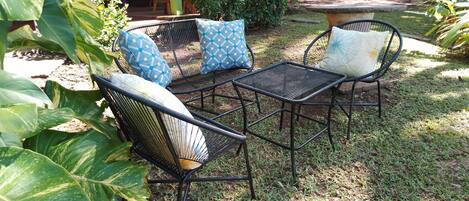 An outdoor seating area with metal chairs and cushions, surrounded by lush greenery and a hammock.