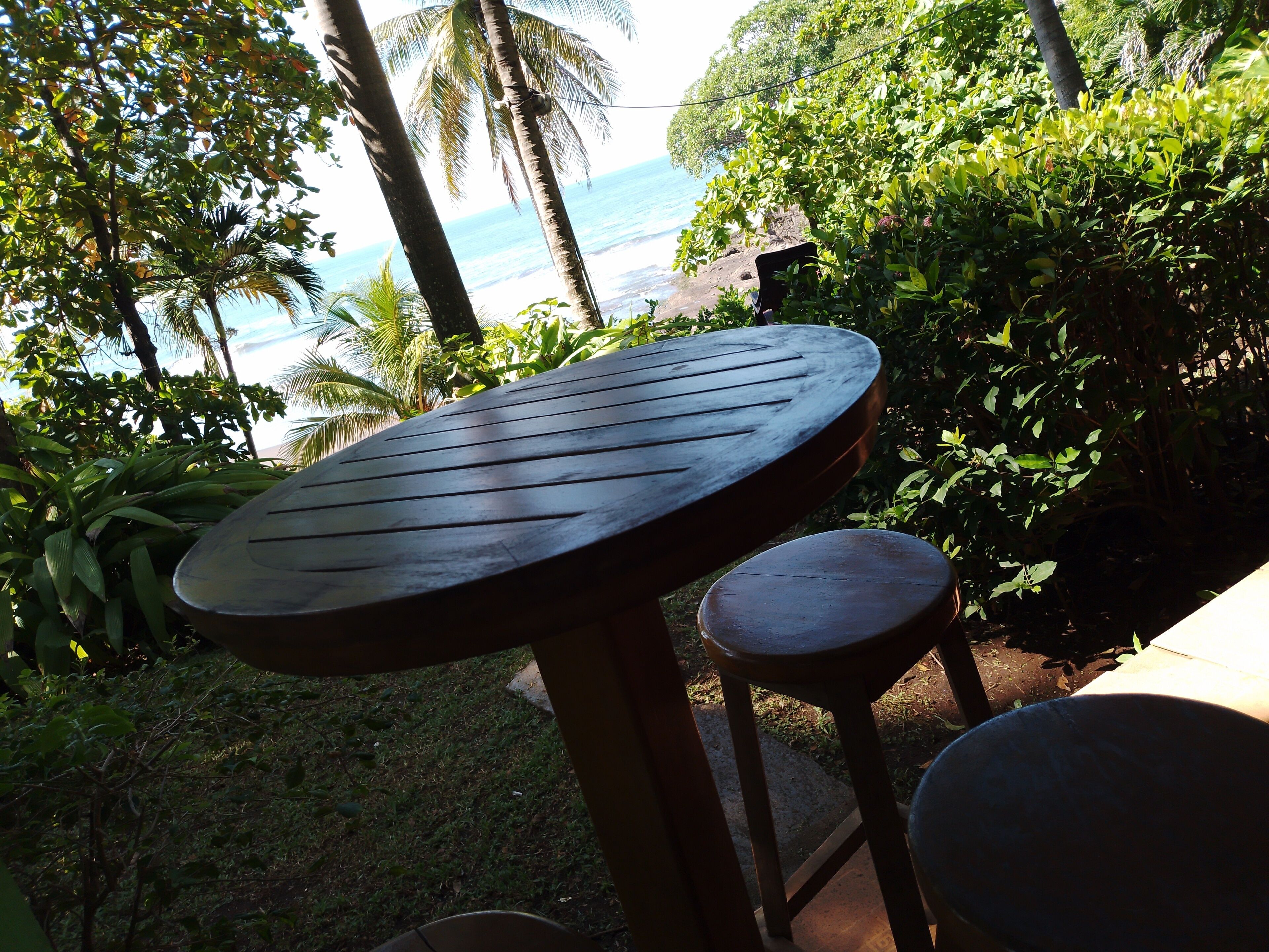 Uma mesa e um banquinho de madeira em um ambiente externo exuberante, com vista para o oceano.