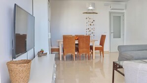 A modern living room with a flat-screen TV, wicker basket, grey sofa, dining table with wicker chairs, and a hanging light fixture. - Apartamento Puerta del Sol (Conil de la Frontera)