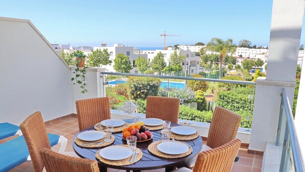 A round table with six wicker chairs, a blue cushion, and a fruit bowl on a balcony overlooking a residential area with a pool and greenery. - Apartamento Puerta del Sol (Conil de la Frontera)