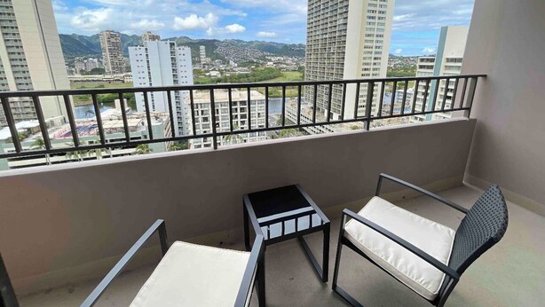 A balcony with a table and two chairs, overlooking a cityscape with tall buildings and mountains. - Heart of Waikiki w/ Kitchen - Free Parking - King Bed - Corner Unit - Mountain View (Honolulu)