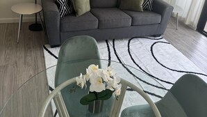 A modern living room with a grey sofa, two black and white patterned throw pillows, a glass coffee table with white flowers, and a black floor lamp. - Heart of Waikiki w/ Kitchen - Free Parking - King Bed - Corner Unit - Mountain View (Honolulu)