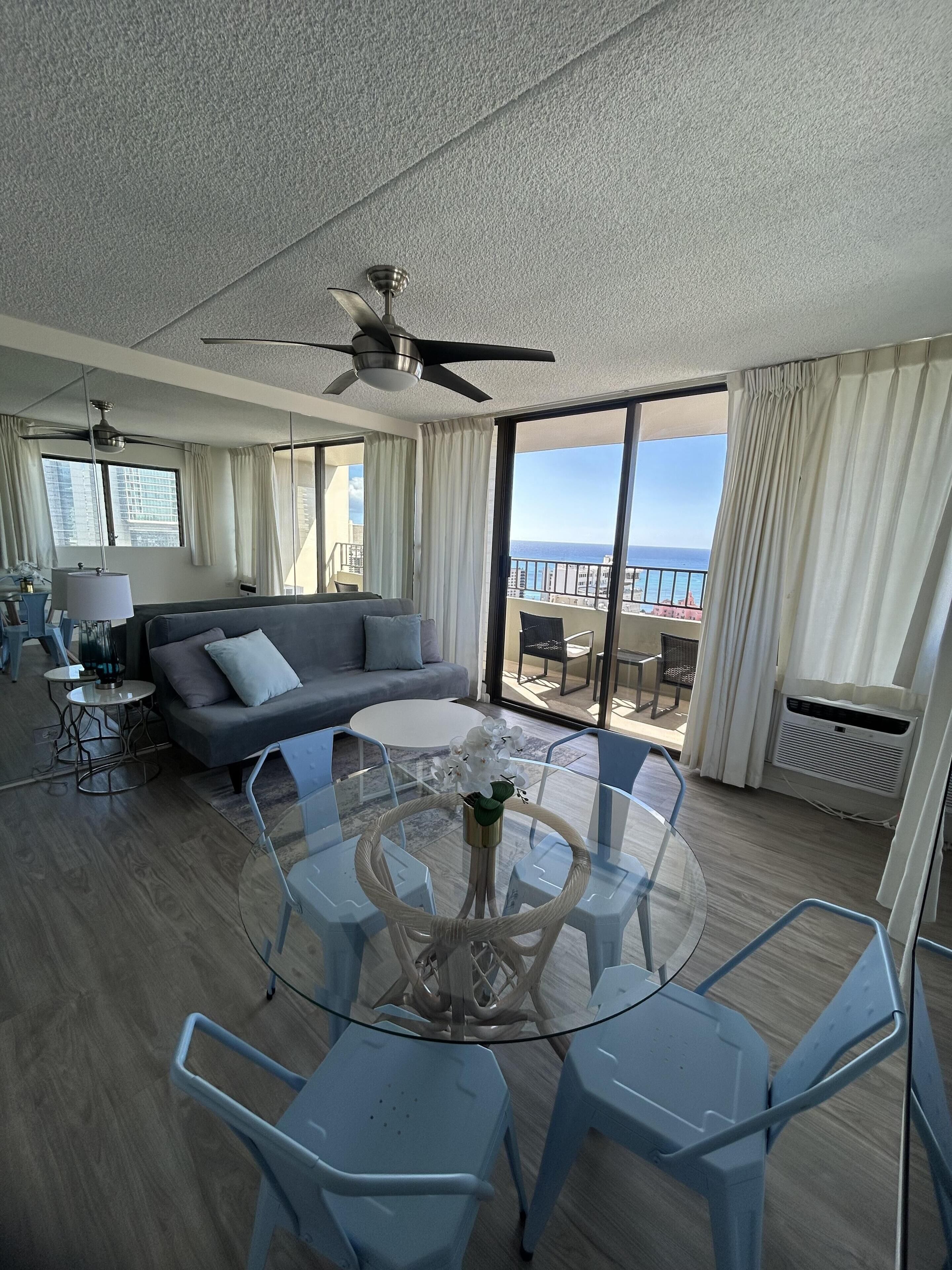 A modern living room with a glass dining table, blue chairs, a gray sofa, and a ceiling fan.
