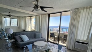 A modern living room with a glass dining table, blue chairs, a gray sofa, and a ceiling fan. - Heart of Waikiki w/ Kitchen - Free Parking - Ocean View - King Bed - Corner Unit (Honolulu)