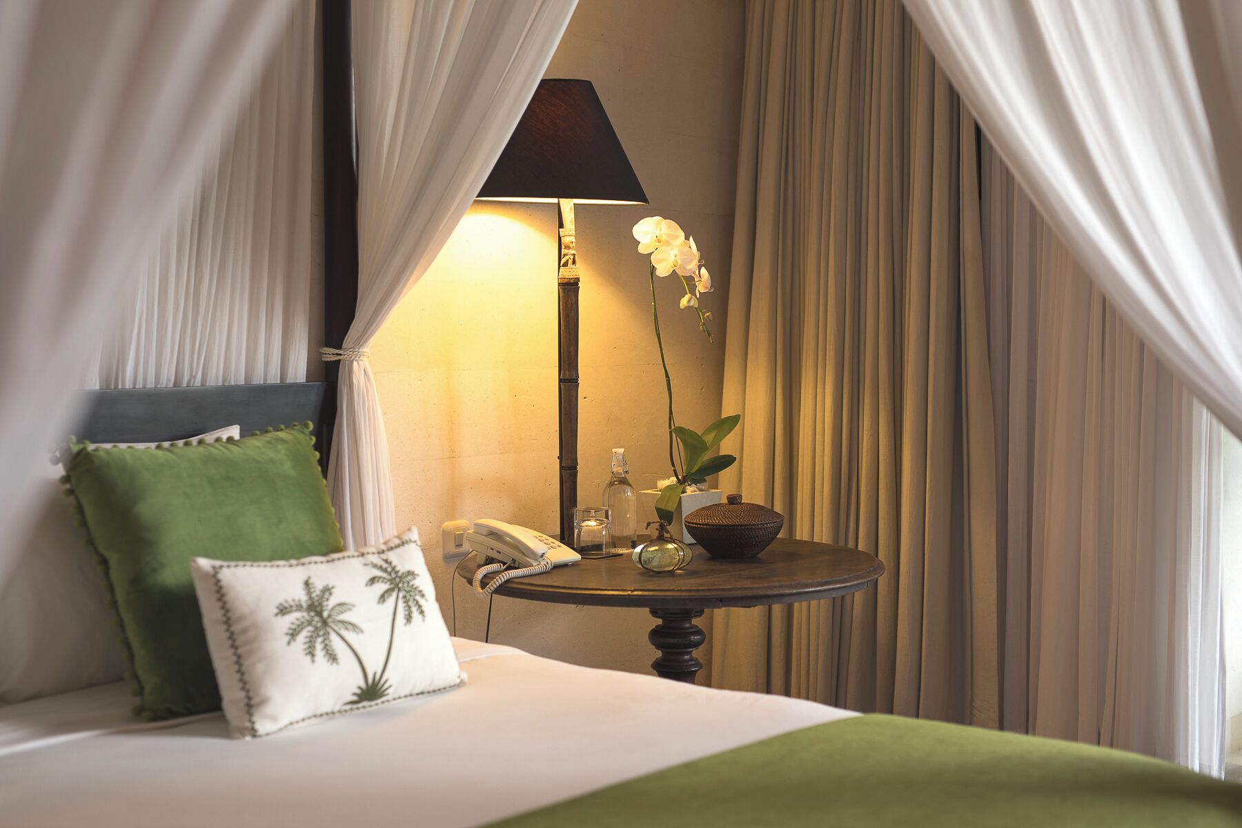 A hotel room with a bed, a bedside table with a lamp, a vase with an orchid, and a green pillow.