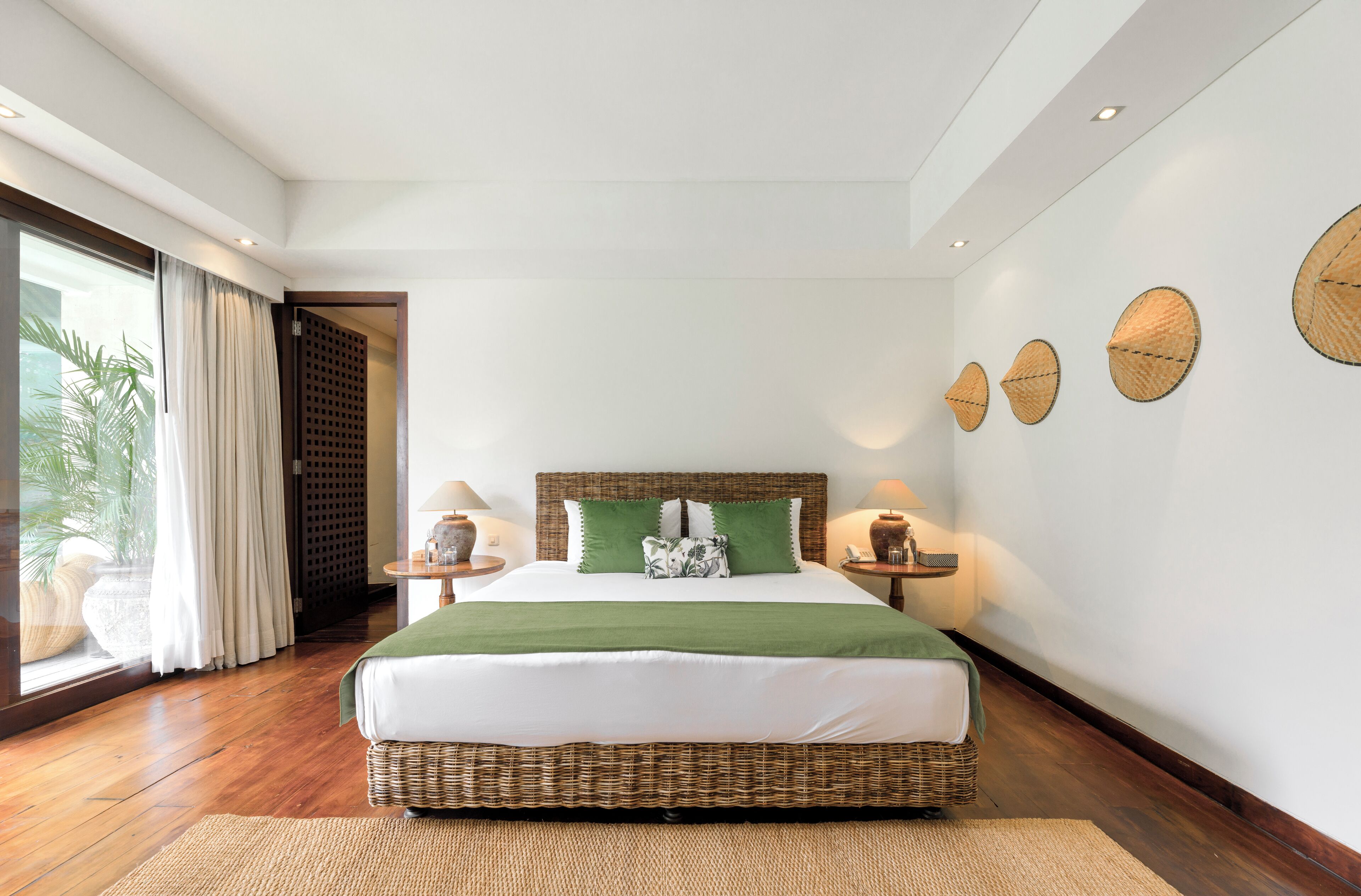 A spacious bedroom with a large bed, a wooden floor, a view of greenery through a window, and a wall with decorative woven items.