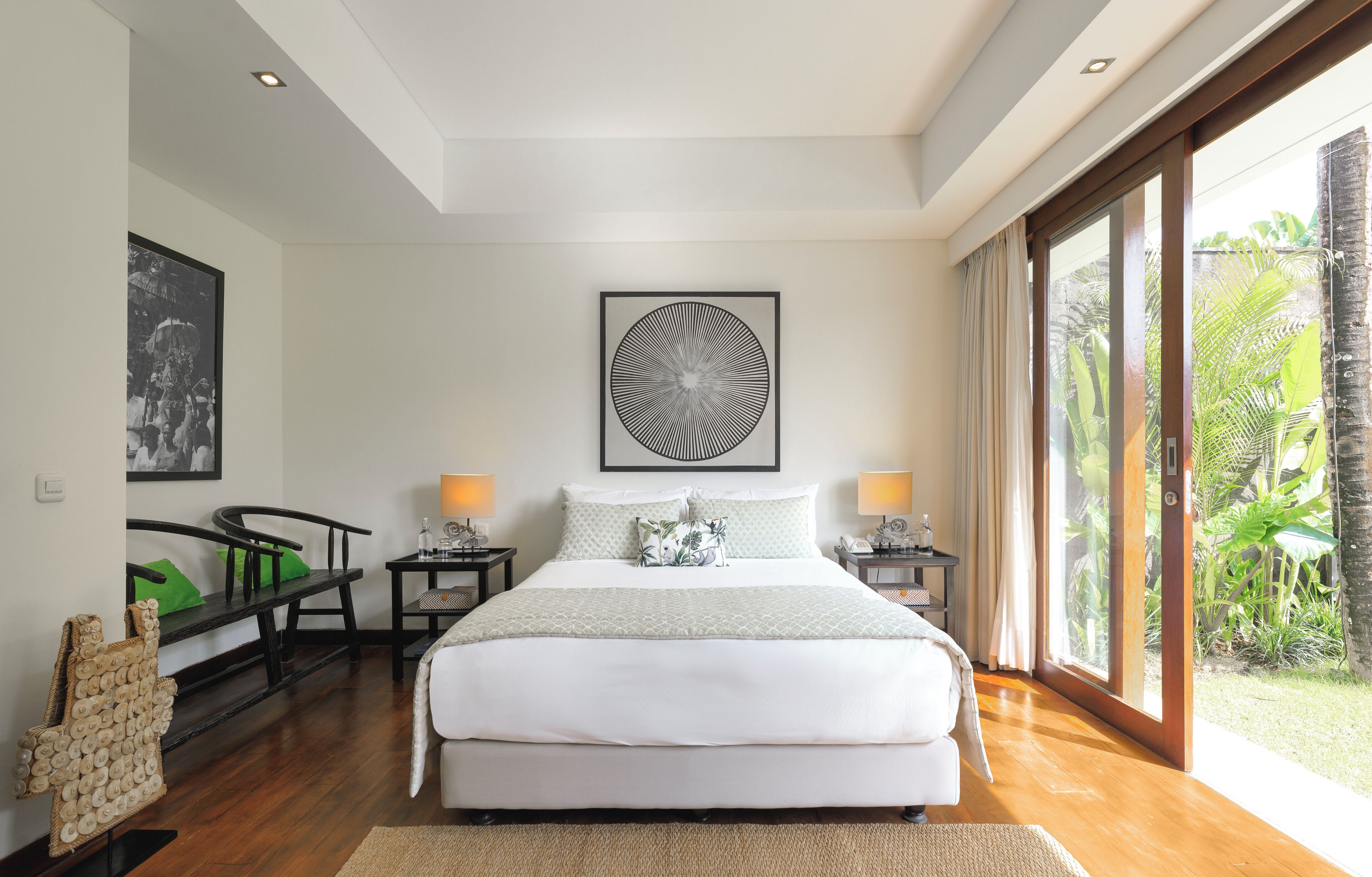 Une chambre spacieuse avec un grand lit, deux tables de chevet avec des lampes, une œuvre d’art encadrée au-dessus du lit et une vue sur la verdure grâce à de grandes fenêtres.