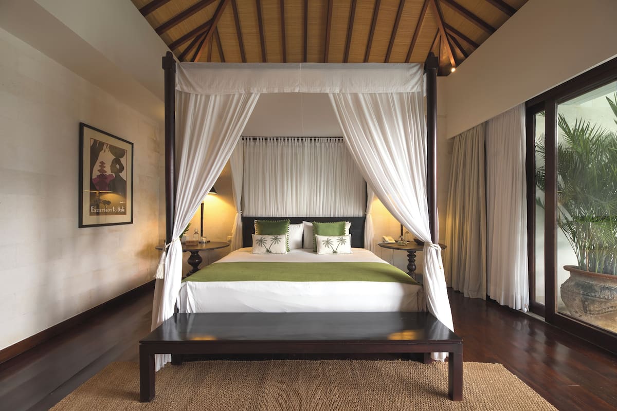 A four-poster canopy bed with white linens and green pillows, a wooden headboard, and a view of palm trees through large windows.