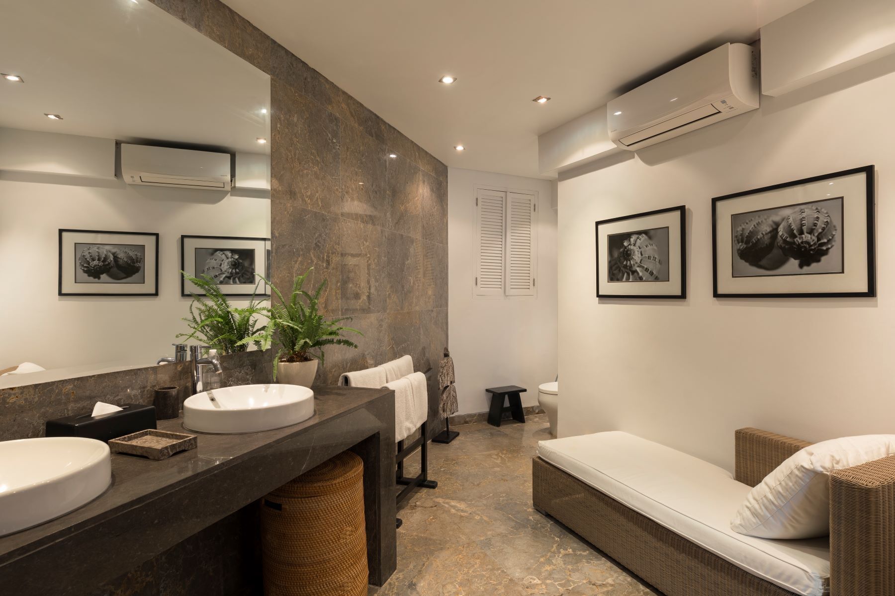 A modern bathroom with a large mirror, two sinks, a toilet, and a wicker sofa.