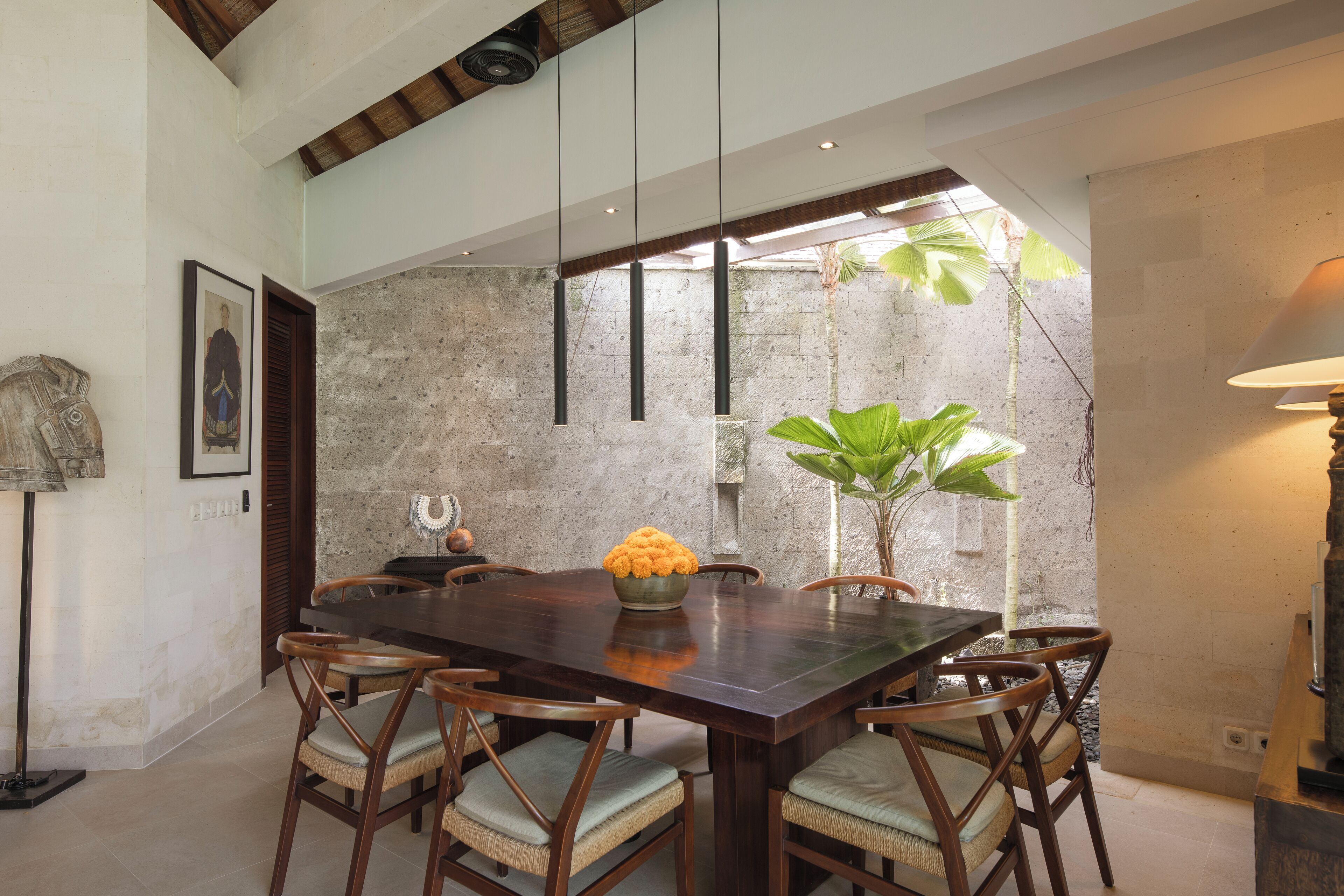 A dining area with a large wooden table, six chairs, a vase with flowers, and a stone wall.