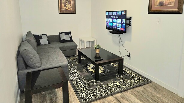 Ein Wohnzimmer mit einem Ecksofa, einem Couchtisch, einem Flachbildfernseher, einem gerahmten Bild an der Wand und einem gemusterten Teppich.