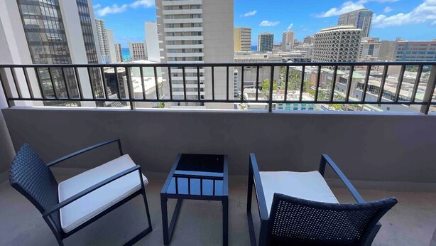 A balcony with two chairs and a small table, overlooking a cityscape with modern buildings. - Heart of Waikiki w/ Kitchen - Free Parking - King Bed - Corner Unit - Partial Ocean View (Honolulu)