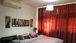 A bedroom with a bed, a wall-mounted air conditioner, a large window with red curtains, and a pendant light. - Serene one bedroom apartment in a quiet residential area, privacy assured  (Accra)