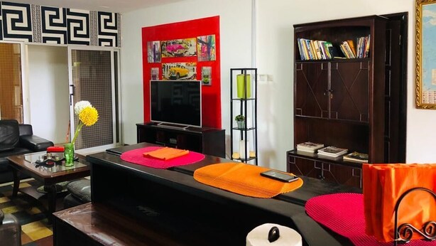 A living room with a black sofa, a coffee table, a red accent wall, a bookshelf, and a ceiling fan. - Serene one bedroom apartment in a quiet residential area, privacy assured  (Accra)