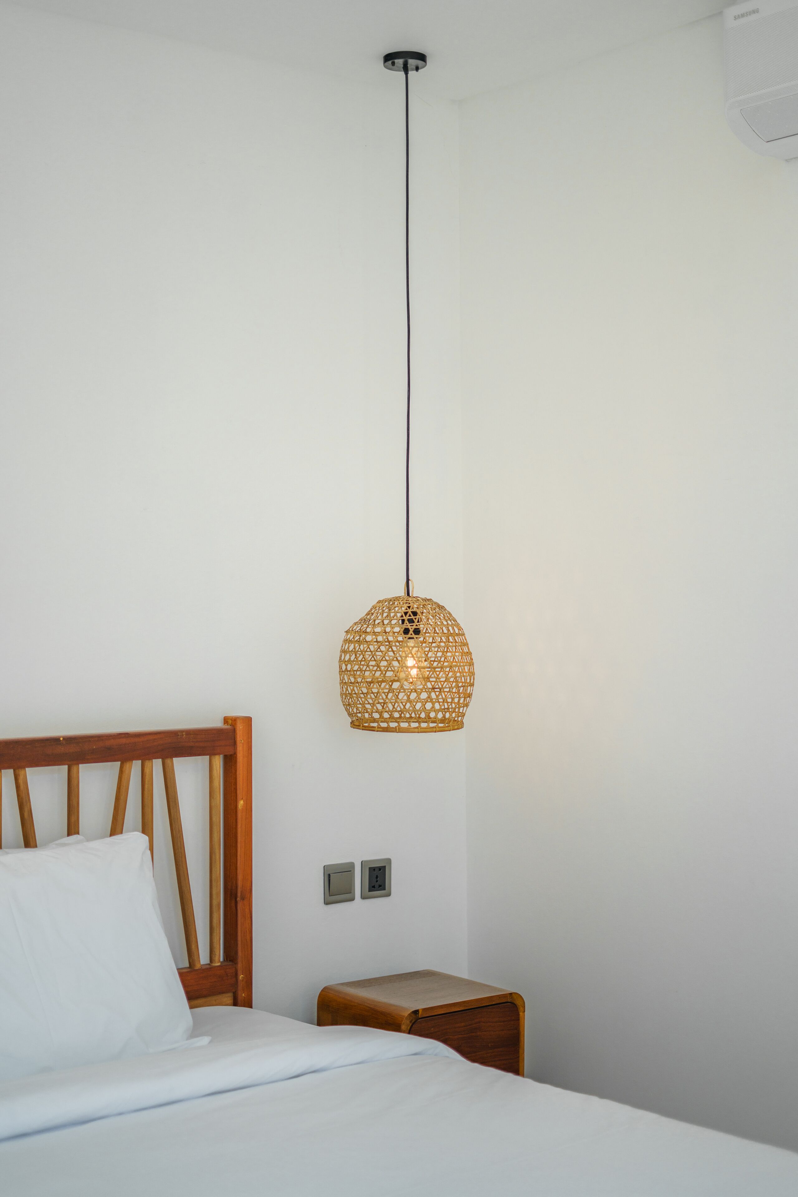 A neatly made bed with a wooden headboard, a small wooden nightstand, and a woven pendant light fixture.