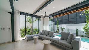 A modern living room with a sectional sofa, two round coffee tables, and a large window overlooking a pool. - Villa Vantage 7 (Kerobokan)