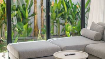 A modern living room with a grey sectional sofa, a round coffee table, and a large window with a view of indoor plants.
