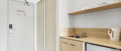 A compact kitchenette with a sink, a small refrigerator, and a shelf with cups and a kettle.