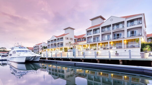 A marina with a white yacht docked, a multi-story hotel with balconies and outdoor seating, and a calm water surface reflecting the buildings. - Hope Harbour Hotel Apartments (Hope Island)