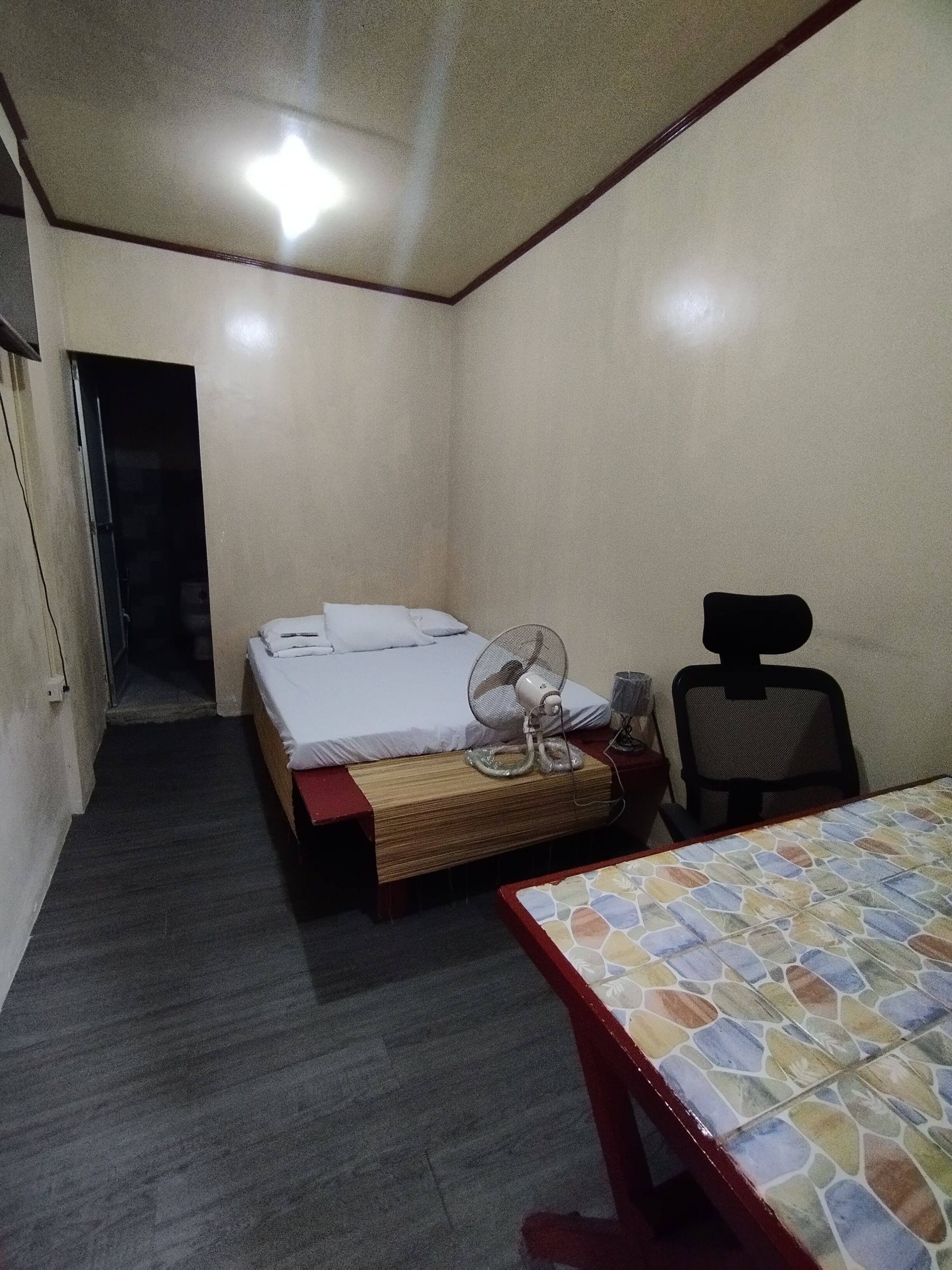 Una habitación pequeña y tenuemente iluminada con una cama individual, una silla, una mesa y un ventilador.