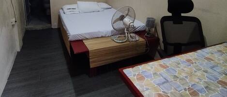 Una habitación pequeña y tenuemente iluminada con una cama individual, una silla, una mesa y un ventilador.