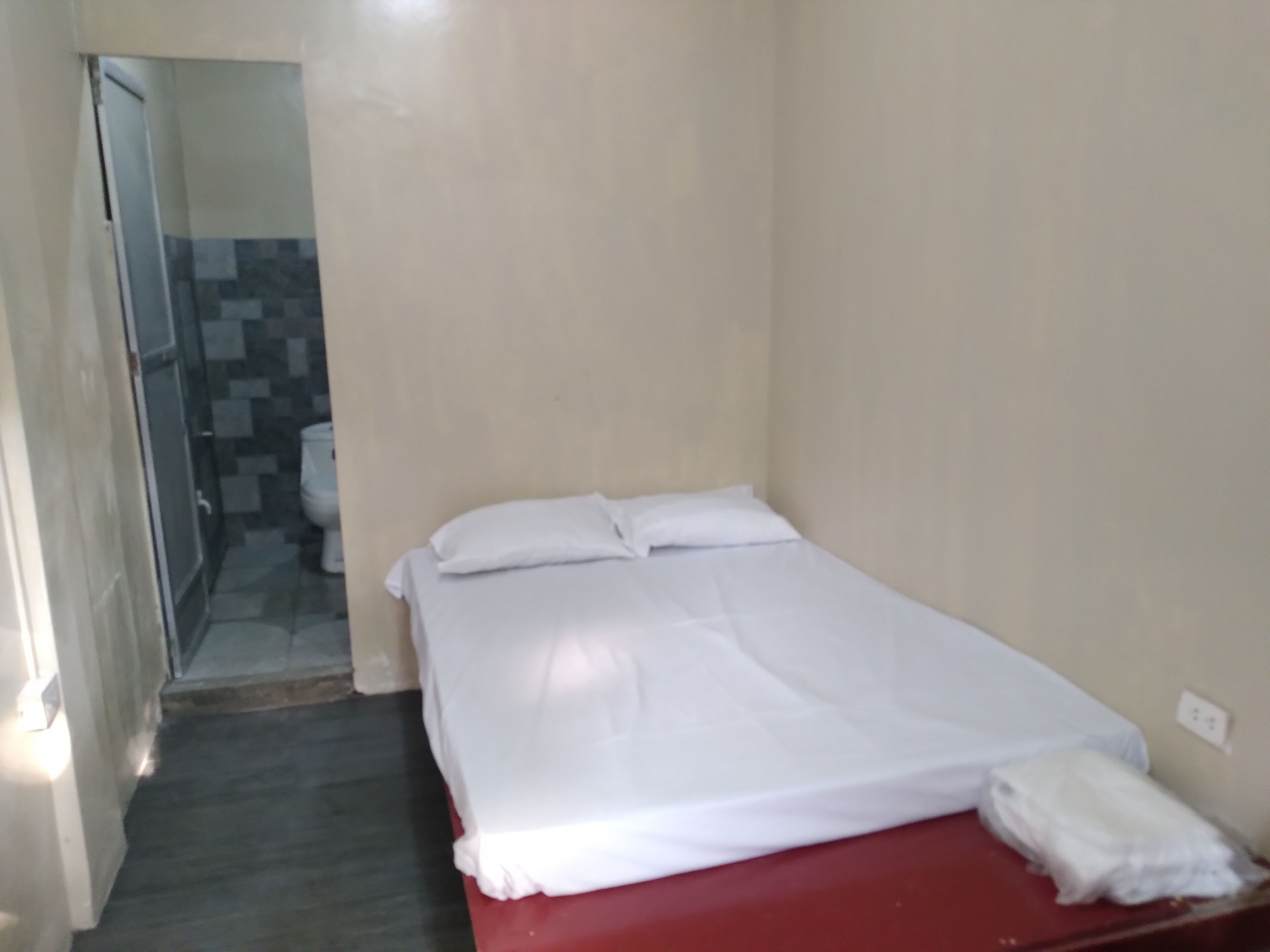 A small, single-bed room with a red bed frame, white bedding, and a tiled shower area visible through a glass door.