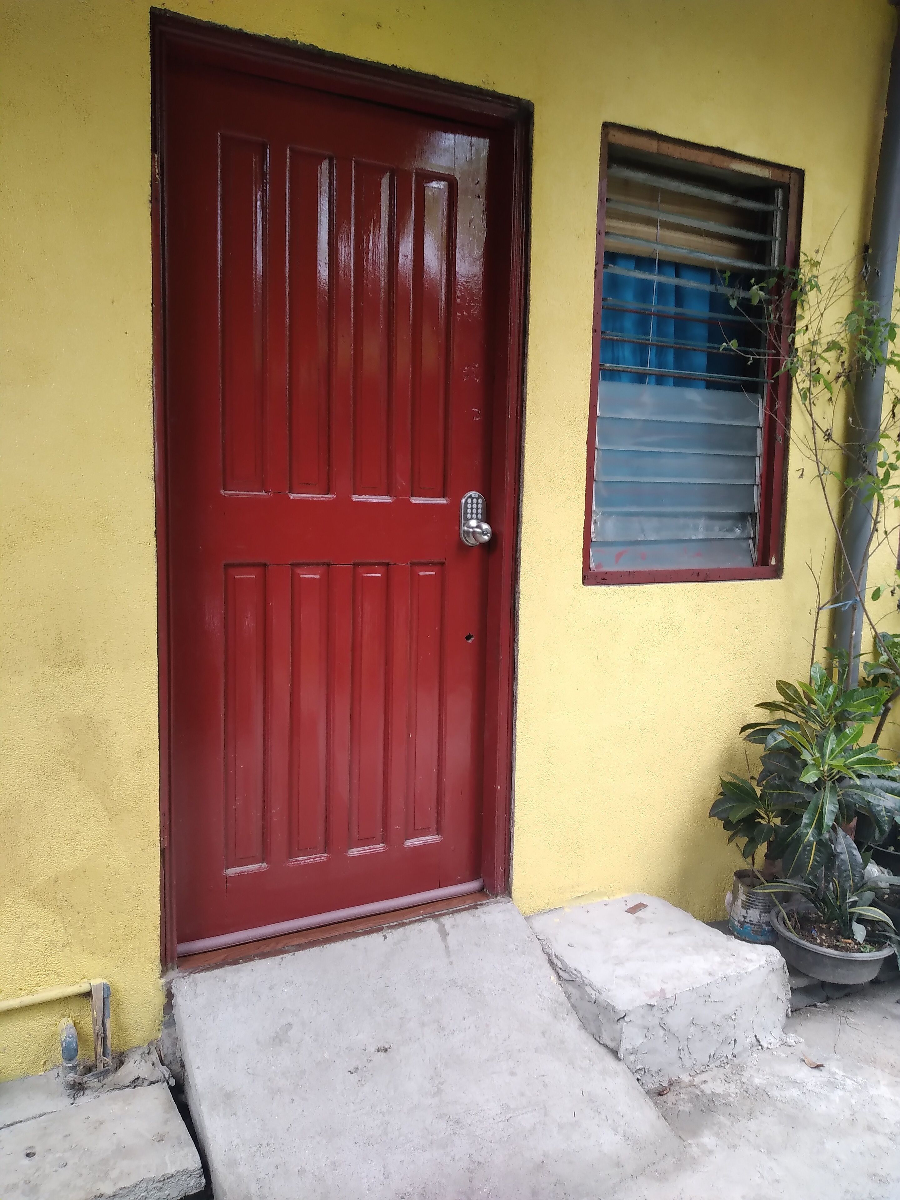 Una puerta roja con cerradura numérica, una ventana pequeña con barrotes y un escalón de concreto que lleva a la puerta.