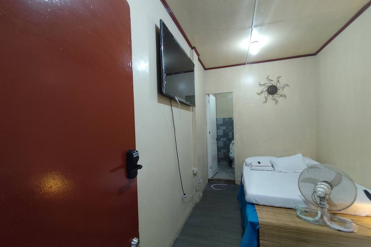 A compact hotel room with a bed, a fan, a TV mounted on the wall, and a door with a security lock.