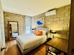 A bedroom with a bed, a desk, a chair, a mirror, and an air conditioning unit.