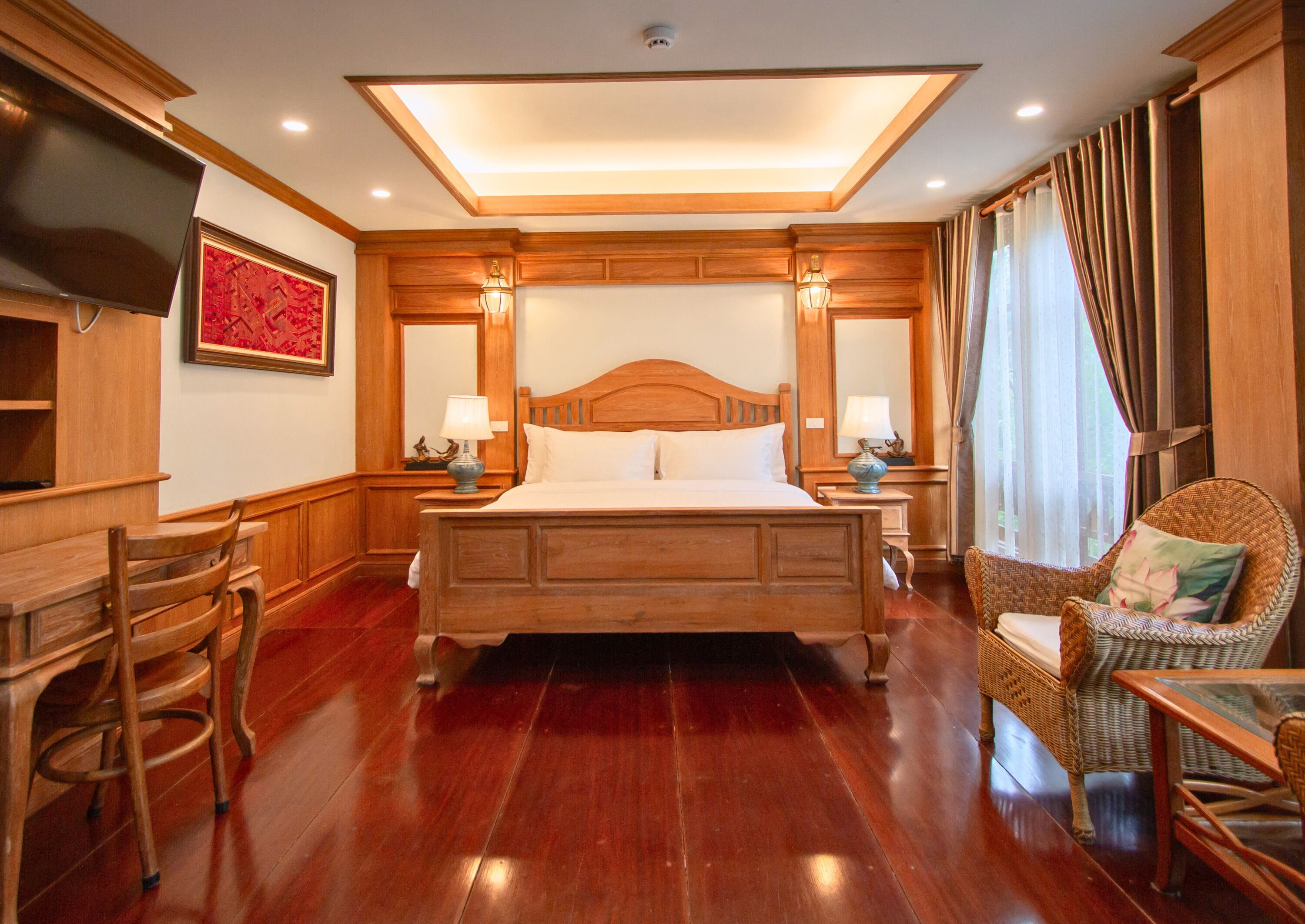 A spacious bedroom with a large wooden bed, two bedside tables with lamps, a wicker chair, and a TV mounted on the wall.
