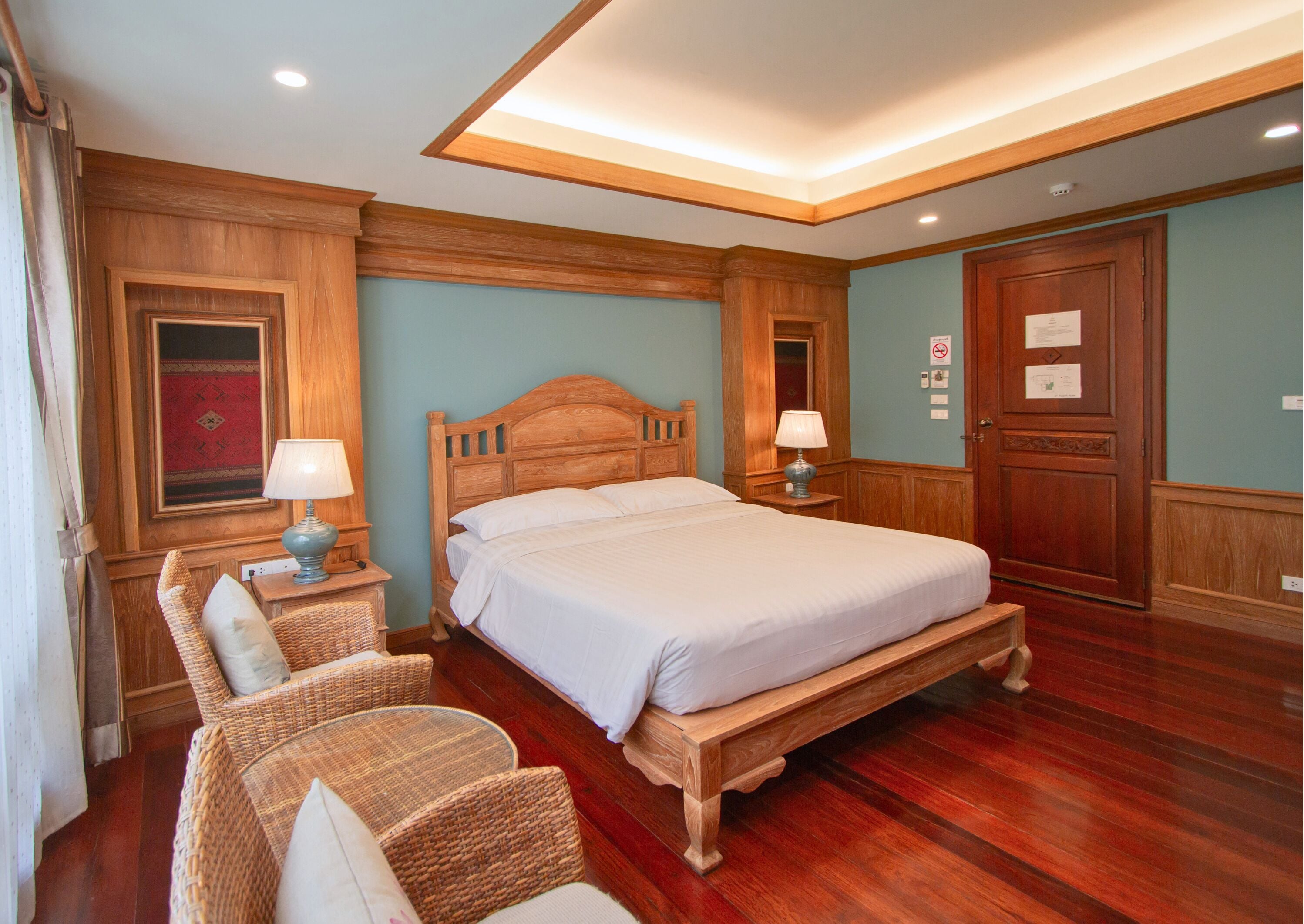 A bedroom with a wooden bed, two bedside tables with lamps, a wooden bench, wicker chairs, and a red patterned wall hanging.